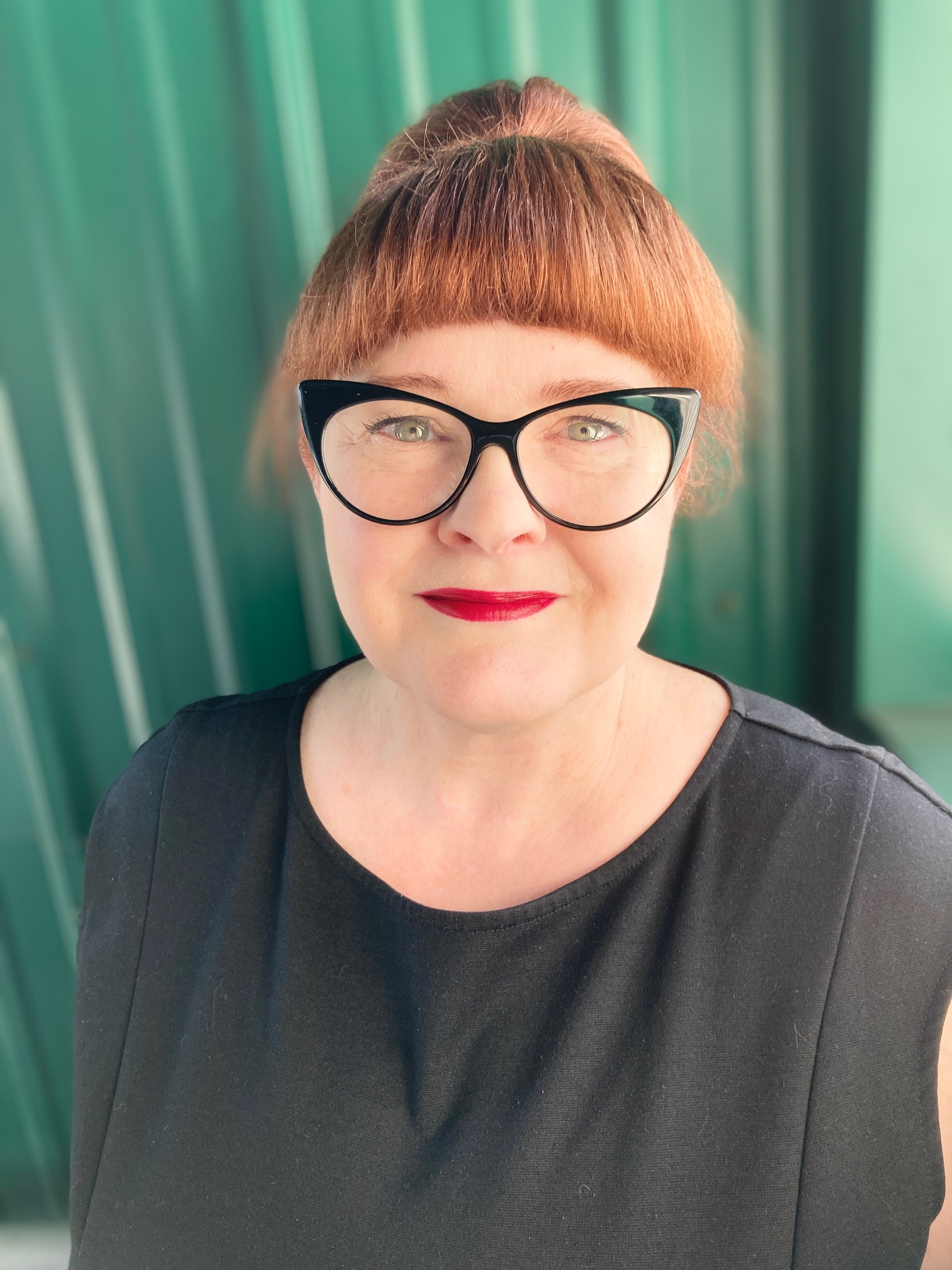 Traci stands in front of a green wall wearing a black shirt and cateye glasses and red lipstick. Her red hair is pulled back from her face.