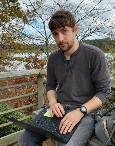 George sits on a bench with his laptop on his lap and a tree in the background.