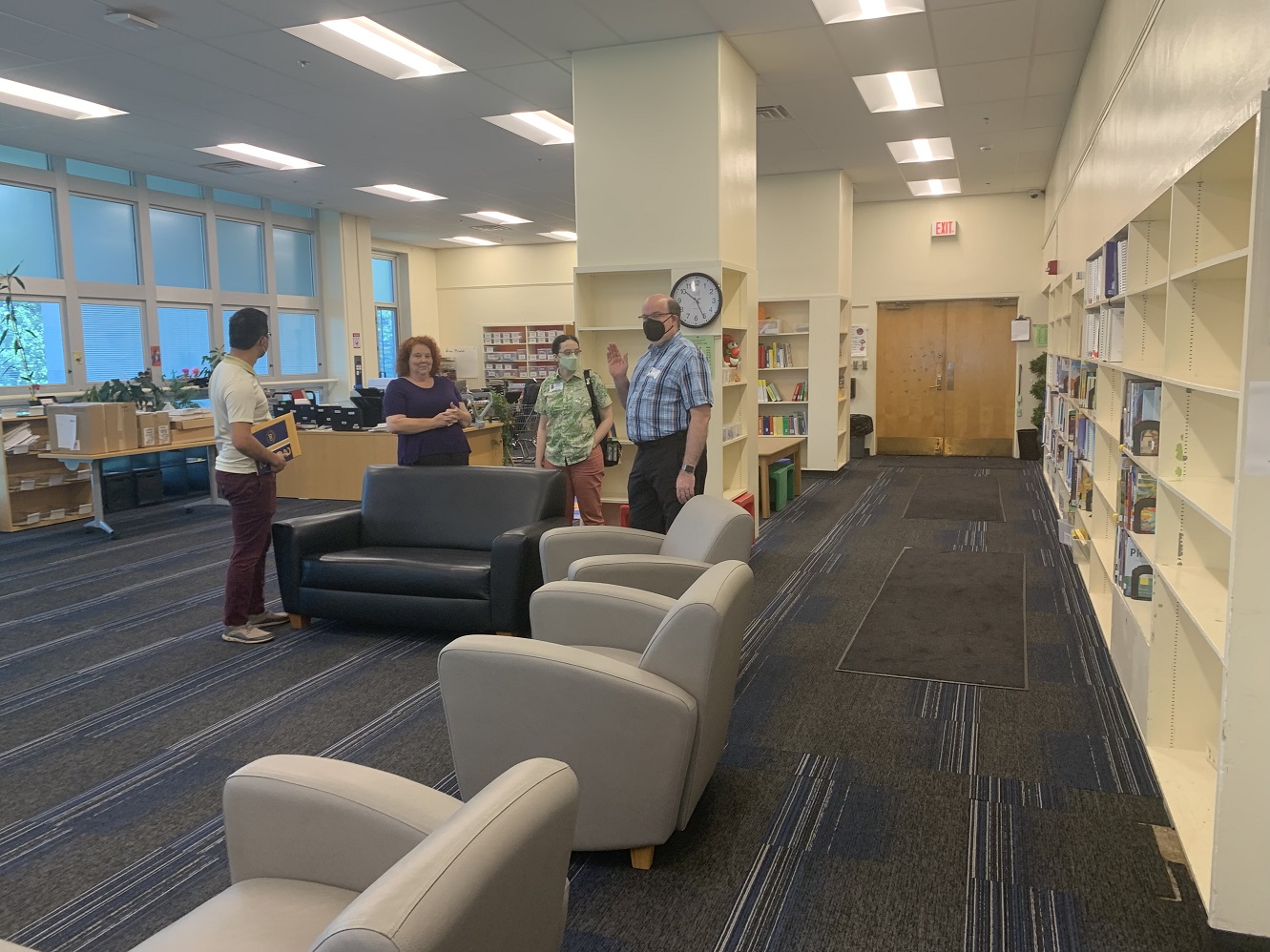 The school librarian is a woman with tightly curled red hair and a purple t-shirt. Beside her is Dr. Hung, Katy, who has braided hair, glasses, a green face mask, and a green shirt with ferns and moths printed on it, and James, who has a black face mask and a blue plaid shirt, and who is waving to the camera. The library has shelves with books and other materials, a large front desk, and an area of comfortable-looking arm chairs and sofas arranged in a semi-circle.