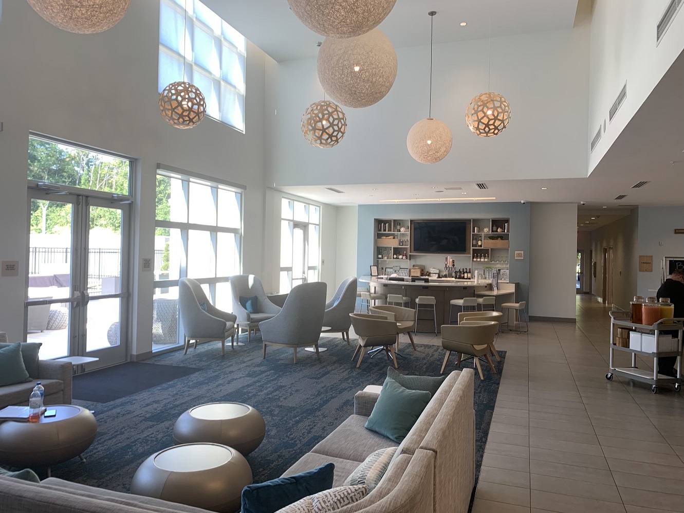 The hotel lobby, showing scattered seating and side tables, and a semicircular bar at the far end. The wall to the left of the viewer is mostly glass, looking out on outdoor seating, a pool, and a lawn. Overhead, round hanging lights contribute to a fun atmosphere.