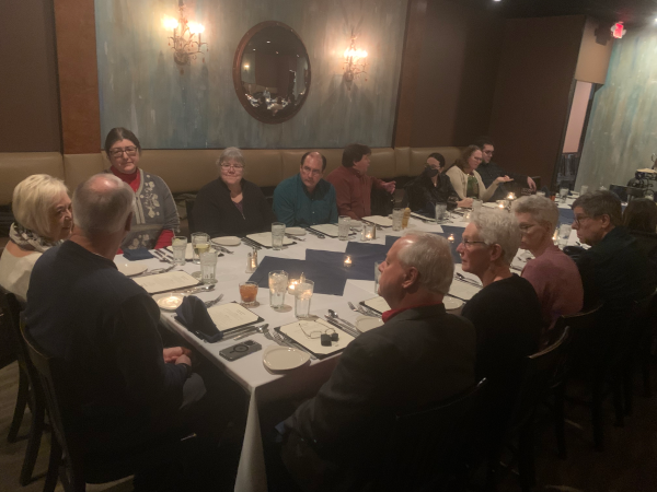 A look at the long table where the Keystone staff are gathered. John and Laura are sitting at the head of the table. On the side facing the camera are Mitake, Nancy, Kyle, Drea's husband, Katy, Marion, and Marion's husband. On the side closer to the camera are Brian with his wife, and Mark with his wife. The room has green textured paint, and a pair of wall lamps beside a convex round mirror.