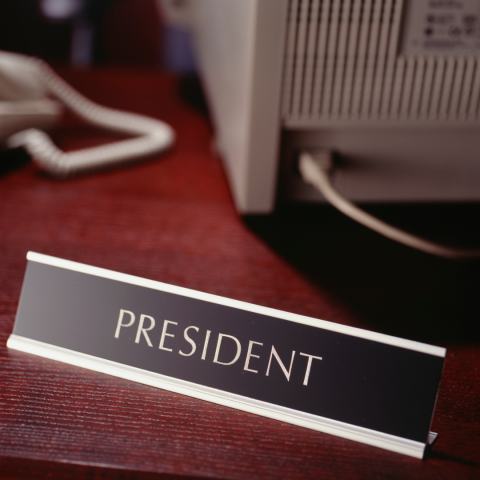 A gold and black "President" title placard sits on a cherry wood desk in front of a telephone and computer.