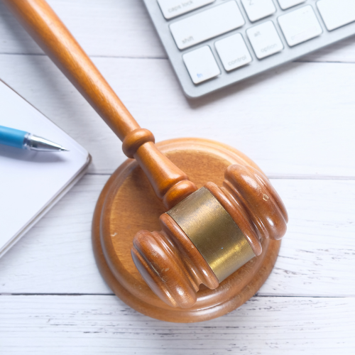 A wooden gavel sits on a table with a laptop, notebook, and pen.